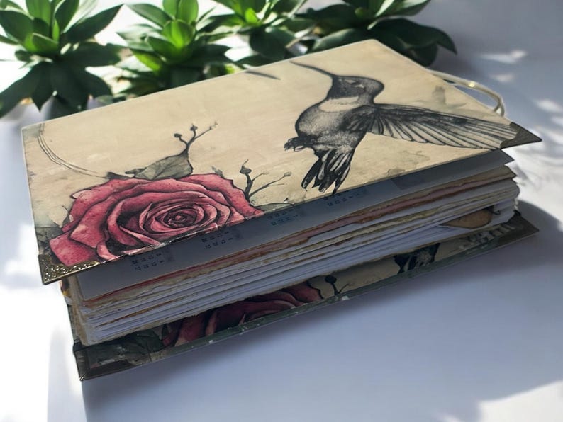 May include: A handmade journal with a beige cover featuring a watercolor illustration of a hummingbird and a rose. The journal has a gold-colored spine and is bound with multiple pages.