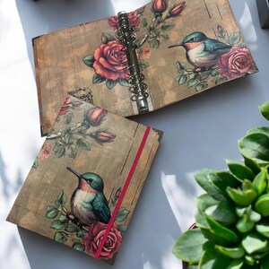 May include: A brown and gold binder with a floral design featuring red roses and hummingbirds. The binder has a red ribbon and a silver ring mechanism.
