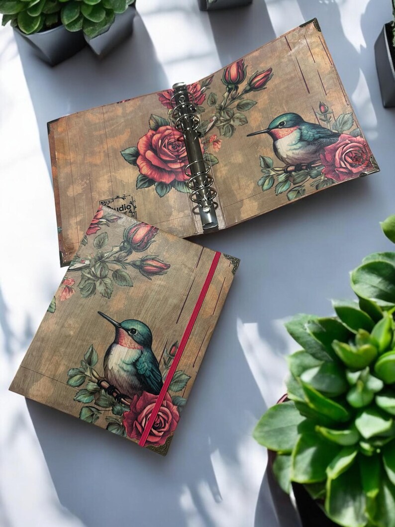 May include: A brown and gold binder with a floral design featuring red roses and hummingbirds. The binder has a red ribbon and a silver ring mechanism.