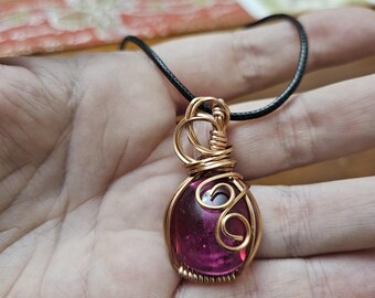 May include: A pink glass gemstone pendant wrapped in gold wire, hanging from a black cord.