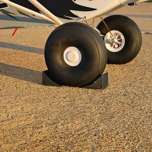 May include: A model airplane with large black tires and a raven graphic on the side. The plane is white and gray with a red accent. The tires are resting on a black stand.