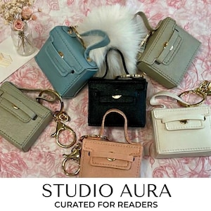 May include: An assortment of miniature handbags in various colors, including blue, black, pink, and white, each with a gold-tone keychain. The bags are arranged on a floral patterned surface. The text "STUDIO AURA CURATED FOR READERS" is at the bottom.