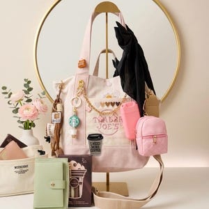 May include: A light pink canvas tote bag with the text Trader Joe's, a daisy patch, and a coffee cup patch with the text I Need More Coffee. The bag is decorated with a gold chain, various charms, a black scarf, and a small quilted pink backpack.