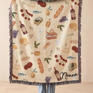 May include: Beige throw blanket with a repeating pattern of food items including bread, cheese, grapes, and wine bottles. The blanket has a fringe border and the word "Nonna" is written on the blanket.