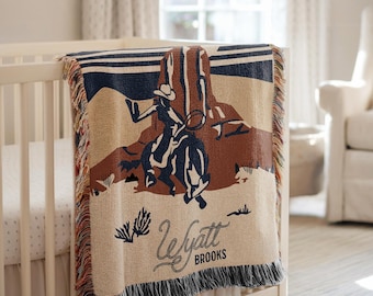 Peut inclure: Une couverture tissée beige et marron avec un motif de cow-boy et de cheval, drapée sur un berceau blanc. La couverture présente un cow-boy attrapant un cheval en bleu marine, avec le nom "Wyatt Brooks" en écriture.