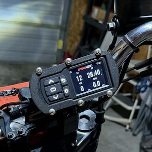 Peut inclure: Un écran numérique Dynojet noir monté sur le guidon d'une moto. L'affichage indique 12 B+, 28.40 RPM et 0.0 VSS. L'appareil est fixé par un cadre noir texturé avec quatre vis argentées et comprend des boutons de navigation intégrés.