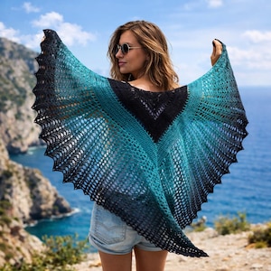 May include: A knitted shawl with a gradient of colors, transitioning from black to teal and turquoise. The shawl has a scalloped edge and is held open against a backdrop of the ocean and sky.