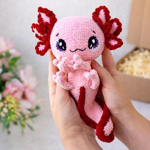 Peut inclure: Une peluche axolotl rose au crochet, faite à la main, avec des branchies et des détails de queue rouges. L'axolotl a de grands yeux expressifs avec des iris violets. La peluche est tenue dans deux mains. Une création charmante et fantaisiste.