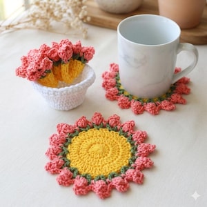 May include: Handmade crochet coasters in a floral design. The coasters feature a yellow center, green trim, and pink flower petals. A white mug sits on one coaster. A small white bowl holds additional coasters.