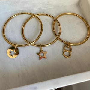 Op de afbeelding: Drie goudkleurige armbanden met verschillende bedels: een stervormige uitsparing, een effen ster en een D-vormig ontwerp. De armbanden worden in een witte schaal gepresenteerd.