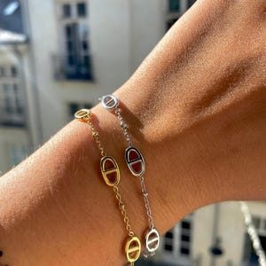 Peut inclure: Deux bracelets, un doré et un argenté, sont portés sur un poignet. Chaque bracelet est composé d'une chaîne avec des maillons ovales. Le bracelet doré a un centre rouge-brun dans les maillons ovales, tandis que le bracelet argenté a un design similaire.