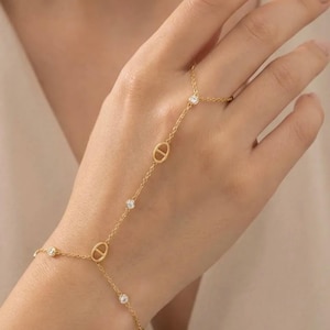 Peut inclure: Une fine chaîne de main en or avec un design de bague, de chaîne et de bracelet. La chaîne est ornée de petites pierres précieuses claires scintillantes et de maillons ovales en or. Le bijou est présenté sur une main.