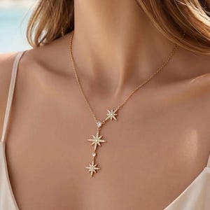 Puede incluir: Un collar de tono dorado con una cadena delicada y tres colgantes en forma de estrella dispuestos en un diseño vertical. Cada estrella está incrustada con pequeñas piedras claras, y dos piedras redondas claras se sitúan entre los motivos.