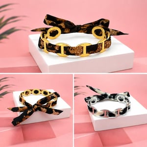 Può includere: Tre braccialetti con un motivo leopardato. I braccialetti hanno dettagli in metallo color oro e un fiocco. Lo sfondo è rosa e bianco.