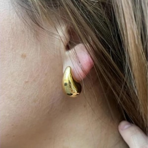 Könnte beinhalten: Nahaufnahme eines goldfarbenen, tropfenförmigen Ohrrings. Der Ohrring ist glatt und glänzend und reflektiert das Licht. Der Ohrring wird am Ohr einer Person mit hellbraunem Haar getragen.