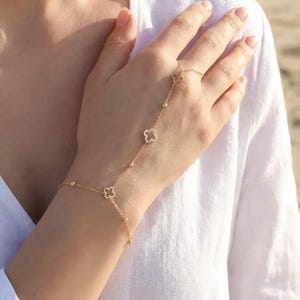 Op de afbeelding: Een delicate gouden handketting met klavervormige bedels en kleine kralen. De ketting verbindt met een ring en wikkelt zich om de hand, waardoor een elegant accessoire ontstaat. De sieraden worden op een hand gedragen.
