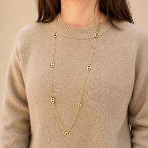 Può includere: Una lunga collana a catena color oro con maglie ovali distanziate uniformemente, ciascuna caratterizzata da una barra orizzontale al centro. La collana è indossata sopra un maglione in maglia beige, mostrando un design delicato e uniforme.
