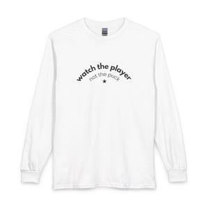 Op de afbeelding: Een wit shirt met lange mouwen en zwarte tekst op de borst met de tekst watch the player not the puck, inclusief een klein zwart stersymbool onder de tekst. Het shirt heeft een ronde hals en geribbelde boorden aan de mouwen.