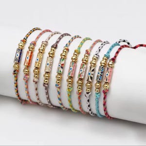 May include: A collection of colorful braided bracelets. Each bracelet features a gold-colored central bead with an eye design in various colors. The bracelets are displayed on a white surface, showcasing their vibrant hues and intricate details.
