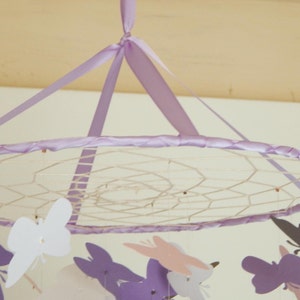 May include: A purple and white mobile with butterflies hanging from a circular frame. The frame is made of a white mesh material and is decorated with a purple ribbon.