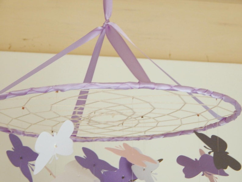 May include: A purple and white mobile with butterflies hanging from a circular frame. The frame is made of a white mesh material and is decorated with a purple ribbon.