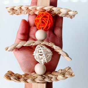 May include: A natural woven bird toy with three tiers. Each tier has a woven ring and a wooden bead. The top tier has an orange ball, the middle tier has a white ball, and the bottom tier has a wooden bead. The toy is attached to a wooden block with a metal hook.