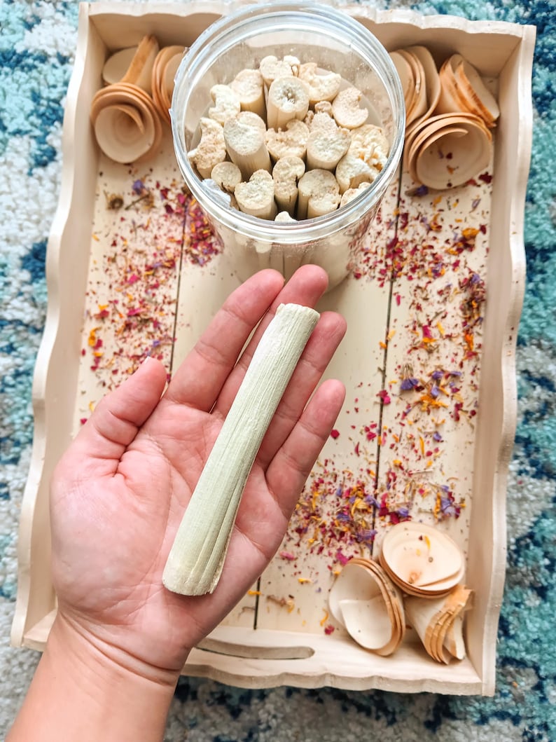 Pu&ograve; includere: Un vassoio di legno chiaro contiene un barattolo di vetro trasparente pieno di piccoli pezzi di legno cilindrici. Una mano tiene un singolo pezzo di legno strutturato, verde chiaro. Piccoli cucchiai di legno e petali di fiori secchi sono sparsi.