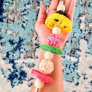 May include: A colorful bird toy with a variety of textures and shapes. It features a yellow loofah, a pink woven ball, a green loofah, a white woven ball, a pink loofah, and wooden beads.