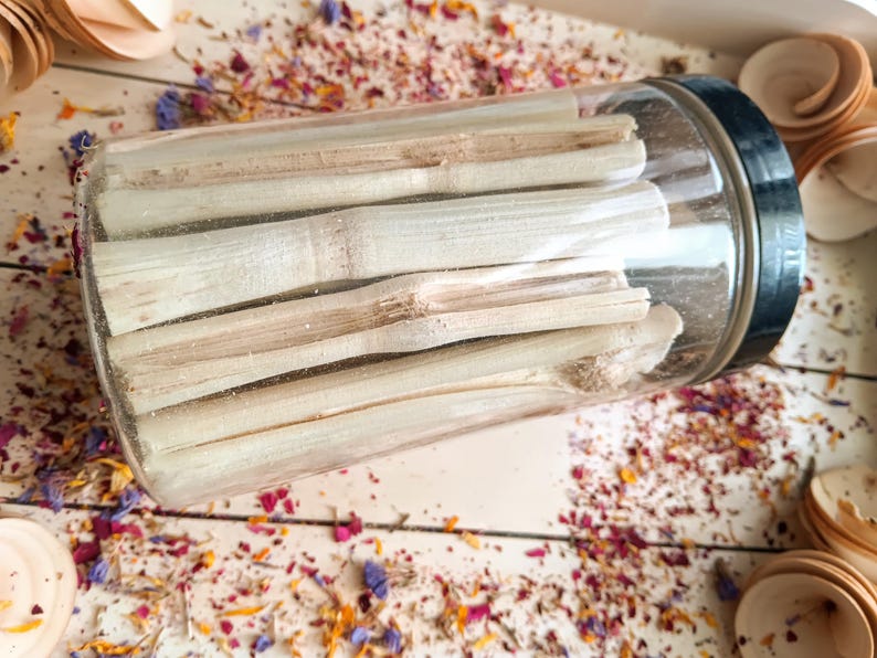 Pu&ograve; includere: Un barattolo di vetro trasparente con coperchio nero pieno di bastoncini di bamb&ugrave; colorati. Il barattolo &egrave; appoggiato su una superficie di legno bianco cosparsa di petali di fiori secchi e piccoli cucchiai di legno. I bastoncini di bamb&ugrave; sono cilindrici.