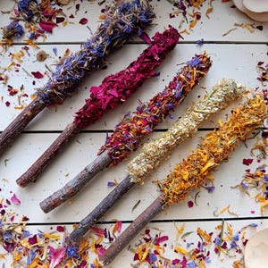 Op de afbeelding: Vijf houten stokjes met verschillende gedroogde bloemenarrangementen. De stokjes zijn bedekt met paarse, roze, rode, gele en witte bloemen.