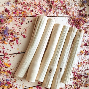 Pu&ograve; includere: Una collezione di sei bastoncini di legno chiari e strutturati disposti diagonalmente su una superficie di legno bianco. I bastoncini sono circondati da una dispersione di petali di fiori secchi nelle tonalit&agrave; del rosa, del viola, del giallo e dell'arancione.