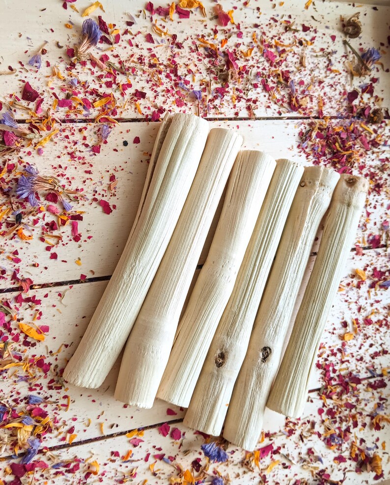 Pu&ograve; includere: Una collezione di sei bastoncini di legno chiari e strutturati disposti diagonalmente su una superficie di legno bianco. I bastoncini sono circondati da una dispersione di petali di fiori secchi nelle tonalit&agrave; del rosa, del viola, del giallo e dell'arancione.