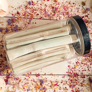Pu&ograve; includere: Un barattolo di plastica trasparente con coperchio nero pieno di bastoncini chiari. I bastoncini sembrano essere steli di piante essiccate. Il barattolo &egrave; su una superficie bianca cosparsa di petali di fiori secchi nei toni del rosa, viola e giallo.