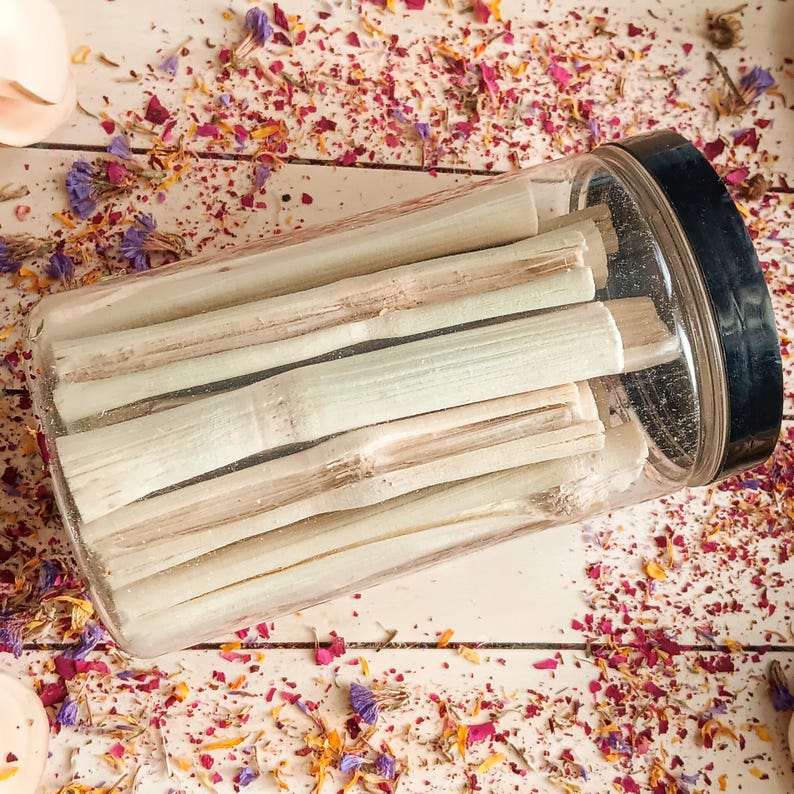 Pu&ograve; includere: Un barattolo di plastica trasparente con coperchio nero pieno di bastoncini chiari. I bastoncini sembrano essere steli di piante essiccate. Il barattolo &egrave; su una superficie bianca cosparsa di petali di fiori secchi nei toni del rosa, viola e giallo.