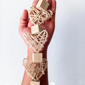 May include: A wooden toy for small pets, featuring four heart-shaped woven pieces strung together with a metal wire. Each heart is attached to a wooden cube.