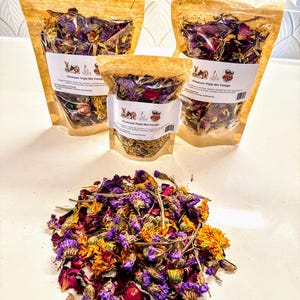 Op de afbeelding: Drie zakjes gedroogde bloemblaadjes en kruiden, gelabeld "Premium Triple Mix Forage", voor kleine dieren. De zakjes zijn gemaakt van doorzichtig plastic met een bruine papieren buitenkant. De bloemen zijn paars, rood en geel.