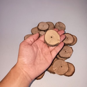 Puede incluir: Un montón de rodajas de madera redondas con un agujero en el centro. Las rodajas de madera son de color marrón claro y tienen un borde de corteza natural.
