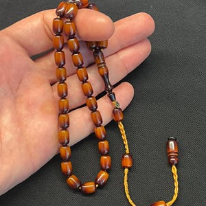 May include: A set of prayer beads featuring oval, amber-toned beads. The beads are connected by a gold-coloured cord. The beads are held in a hand against a dark backdrop.