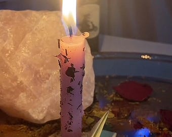 May include: A purple candle with black markings is lit and burning. The candle is sitting on a table with a pink rock and green leaves. There is a small, folded, light green piece of paper in front of the candle.