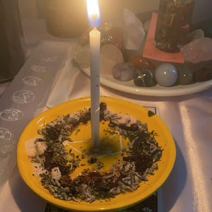 May include: A white candle burning on a yellow plate with herbs and crystals. The plate is on top of a tarot card that reads "Ace of Pentacles".