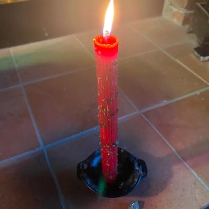 May include: A red candle with a flame burning at the top, sitting in a black metal candle holder. There are three small stones next to the candle holder, one white, one red, and one brown.