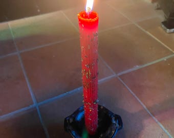 May include: A red candle with a flame burning at the top, sitting in a black metal candle holder. There are three small stones next to the candle holder, one white, one red, and one brown.