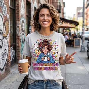 Peut inclure: T-shirt gris clair avec l'inscription "OVERSTIMULATED & UNDERDRESSED" et un dessin animé coloré d'une personne stressée tenant une tasse. La personne sur l'image sourit et tient une tasse de café.