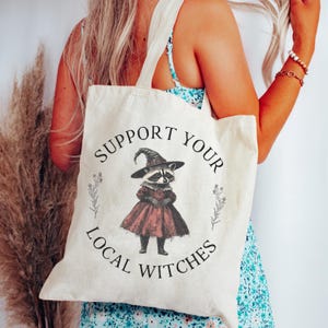 Peut inclure: Un sac fourre-tout en toile blanche avec le texte "Support Your Local Witches" imprimé en noir. Un dessin animé de raton laveur portant un chapeau de sorcière et une robe rouge est au centre du texte.