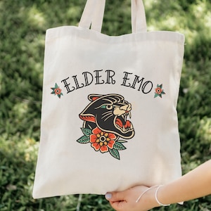 May include: A white canvas tote bag with black handles. The bag features a black panther head with a red and orange flower design and the text "ELDER EMO" printed on it.