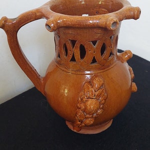 Puede incluir: Una jarra de cerámica marrón con un diseño único. La jarra tiene una parte superior ancha y abierta con un borde decorativo calado. El cuerpo de la jarra está decorado con un diseño floral y tiene una pequeña asa.