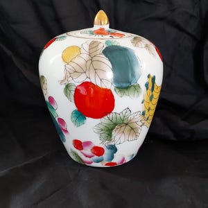 May include: A white ceramic jar with a lid, adorned with colorful hand-painted fruit and leaf designs. The jar features a gold-colored finial on the lid and is set against a dark background. The fruit includes red apples, yellow lemons, and green leaves.