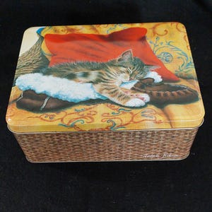 May include: A rectangular metal tin with a woven basket design on the sides. The lid features a painting of a sleeping kitten on a pillow and a pair of moccasins. The kitten is brown, gray, and white.
