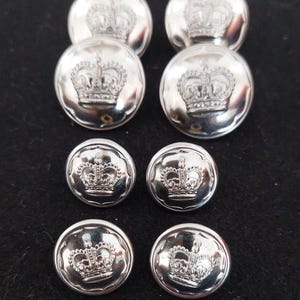 May include: A set of six round, silver-coloured metal buttons arranged in three rows. Each button features a polished, reflective surface with a raised, embossed crown design centred on the face. The buttons vary in size, with two larger ones at the top.