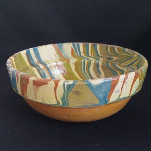 Puede incluir: Un cuenco de cerámica con un borde ancho y una base redondeada. El cuenco presenta un diseño veteado en tonos azules, verdes, marrones y crema. La base es de color terracota sólido, creando un contraste con el interior estampado.